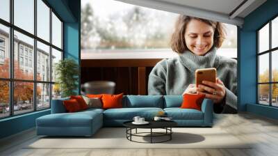 Young woman in a green sweater using a mobile phone by the window in her country house with a view of the mountains and pine trees chalet Wall mural