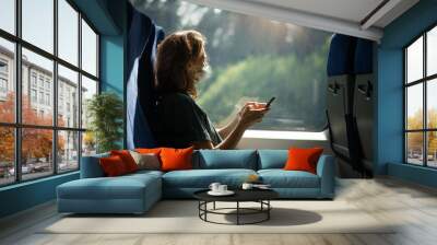 Young woman in a protective mask sitting in a train with a smartphone in her hands. Taking care of health and travel during a pandemic Wall mural