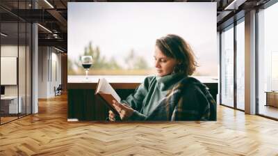 Young woman in green sweater reads a book and drinking wine near window in country house Wall mural