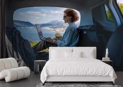 Young woman sitting in the open trunk of a car overlooking the sea and using a laptop, freelancer, remote work and summer vacation auto travel Wall mural