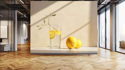  a glass of lemon water next to two lemons on a white table with a shadow cast on the wall behind it. Wall mural