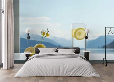  a glass of water and two lemons on a table with a view of mountains and a body of water. Wall mural