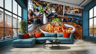  a plate of pasta with tomatoes and basil on a wooden table in a restaurant with people sitting at tables in the background. Wall mural