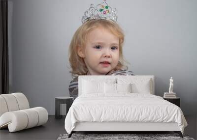 A beautiful baby girl 2 years old in a silver crown and a shiny dress is sitting on a chair on a gray background Wall mural