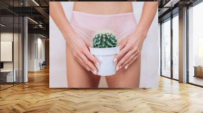 Depilation in the bikini zone concept. A woman holding a cactus in her hand staying in light pink pants with light backghound. Wall mural