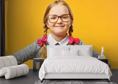 Portrait of a cute little baby girl with glasses on a yellow background. Child schoolgirl made hands in his hips and looks into the camera. The concept of education. Wall mural