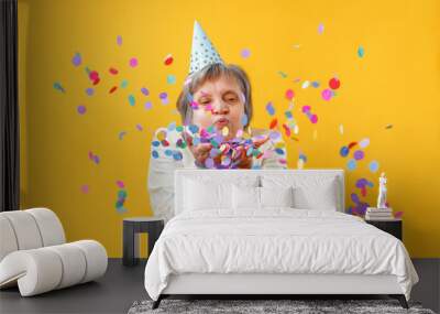 Portrait of a senior woman in studio on a yellow background. Cheerful lady blowing on confetti. Party concept Wall mural