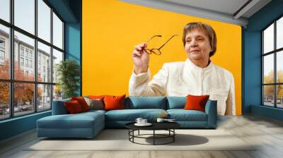 Senior mature charming woman looks in glasses standing on a yellow background Wall mural