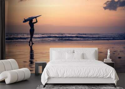 Silhouette of woman walking along the beach with surf board at sunset on tropical island Wall mural