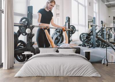 Slim woman in gym with personal trainer exercising power workout with a barbell lying on bench. Wall mural