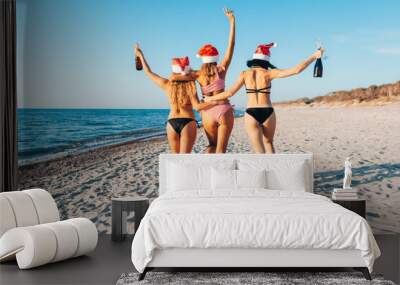 Three young women in bikinis and Santa hats hugging walk on the beach. Women celebrate Christmas and new year at tropics. Back view. Wall mural