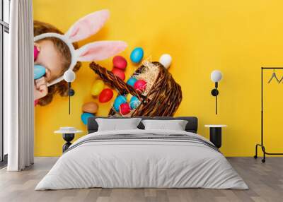 Top view on a happy merry child in bunny ears and a basket with Easter colored eggs. Little girl lies on the floor on a yellow background. Copy space Wall mural