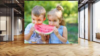 Two kids eating one slice of watermelon in the garden. Kids eat fruit outdoors. Healthy snack for children. 2 years old girl and boy enjoying watermelon. Wall mural