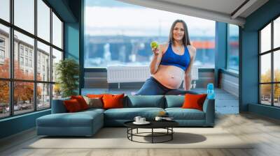 Young happy pregnant woman sitting on yoga mat with green apple and smiling at camera, active future mother living healthy lifestyle eating fruit and drinking water during sport workout at gym Wall mural