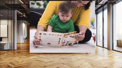 Caring mother showing focused little son book with colorful pictures of living creatures sitting on soft rug in nursery woman educating baby boy at home Wall mural