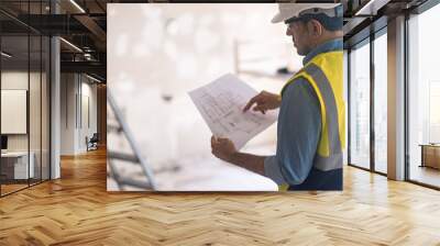 Engineer in professional uniform holding apartment project plan checks details before doing renovation worker in vest holding paper in hands stands against ladder in room Wall mural