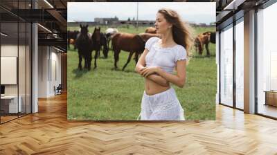 Beautiful girl in a white dress on a background of grazing horses Wall mural