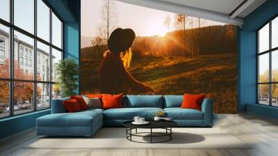 A dreamy young girl or woman traveler in a fedora hat sits in the grass and enjoys the mountain view, the warm rays of the autumn sun and a beautiful sunset. Wall mural