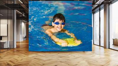 A little boy of 10 years learns to swim in the sport pool using a board. The boy wore swimming goggles. The child is cheerful and happy. Healthy kid enjoying active lifestyle. Wall mural