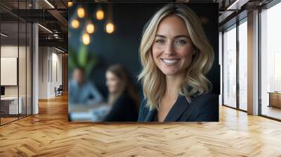 A beautiful blonde woman in her late thirties, smiling and standing at the front of an office meeting room with team members sitting around a table. She is wearing business attire Wall mural