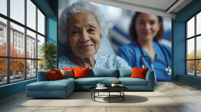 A happy elderly woman in a hospital gown sitting on a chair, next to her is an adult nurse wearing blue scrubs with a stethoscope around their neck, looking at the camera and smili Wall mural