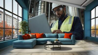 A man in an industrial setting, wearing safety gear and using his laptop to monitor the activity of workers at plants or factories. The background is filled with large steel pipes Wall mural