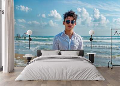 Man in white shirt and sunglasses standing on beach next to the ocean. Wall mural