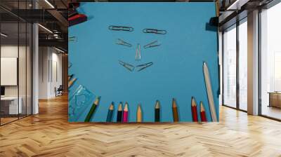 On a blue background are pencils, pen, notebooks, ruler, stapler. With metal clips in the middle is a smiling face. Wall mural