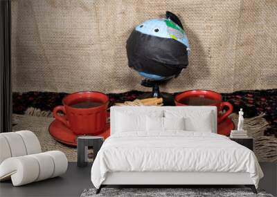 On a jute napkin are two cups of coffee, coffee beans, anise and cinnamon sticks. In the background there is a globe with toy eyes wearing a black medical mask. Wall mural