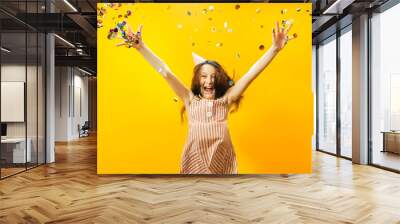 Portrait of a little pretty girl celebrating her birthday. Kid throws colored confetti in both jumps. Yellow studio background. The child is fun and very happy. Wall mural