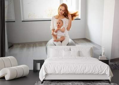 Happy mother holding baby girl while bouncing on fitness ball in cozy home interior. Wall mural