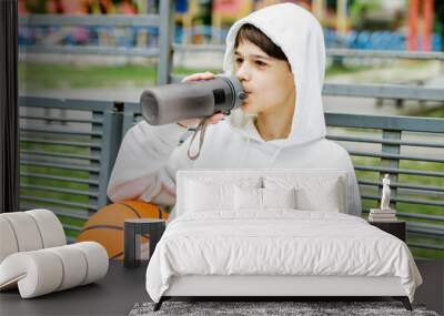 portrait of teenager boy sitting on bench with basketball and drinking water during break Wall mural