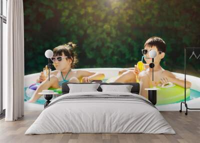 Two young children, boy and girl, sibling, resting in summer in inflatable pool. Children swim in inflatable circles and drink orange juice from tubes. Sunny summer day. Happy summer vacation. Wall mural