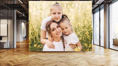 Happy young mother cuddles with her daughters in the summer on the field of rape. Happiness with family Wall mural