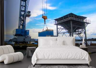 A heavy-duty industrial crane lifting a large blue shipping container at a busy port, set against a backdrop of a bright blue sky and ocean. The scene highlights the efficient cargo handling process Wall mural