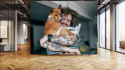 Crazy haired hipster in striped shirt and tattoos hugging dog in industrial loft apartment Wall mural