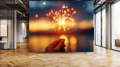 A hand holds a sparkling firework against a colorful sunset, capturing a moment of celebration and joy near water. Wall mural