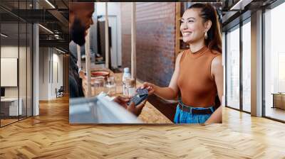 Happy hispanic customer paying for a meal in a restaurant using a nfc machine and credit card. Smiling young woman making a purchase in a store with her debit card and a pos machine. Woman paying bill Wall mural