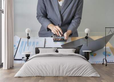 Young Asian business woman sitting at a table holding a pen and using a calculator to calculate graphs and plan the process of growing a successful business Wall mural