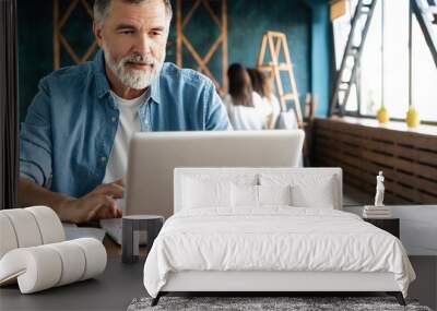 Cheerful mature man working on laptop and smiling while sitting at his working place Wall mural