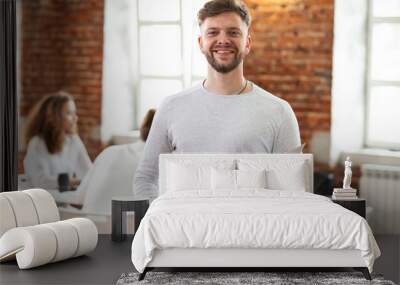 Confident team leader. Confident young man keeping arms crossed and looking at camera with smile while his colleagues working in the background. Wall mural