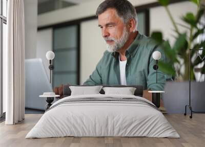 Handsome mature man in casual suit sitting at the table in home office and working at laptop Wall mural