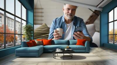 Happy smiling senior man using smartphone device while sitting on sofa at home Wall mural