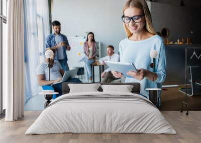 Leading her team to success. Confident young woman holding digital tablet and looking at camera with smile while her colleagues working in the background. Wall mural