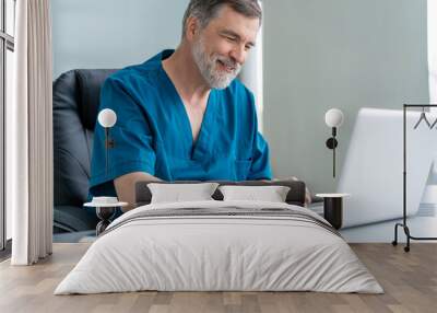 Mature male doctor working on laptop computer, sitting in medical office. Wall mural