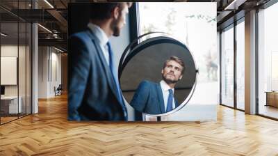 Reflection of handsome young man in full suit standing near the window in front of the mirror indoors Wall mural