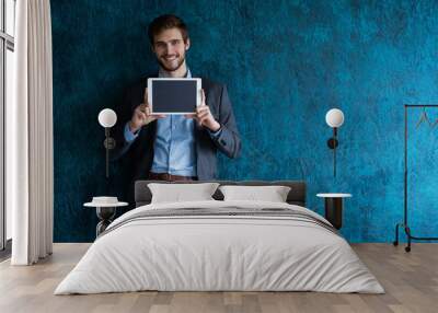 Smiling successful young businessman on blue background in a classic gray suit presenting a tablet. Wall mural