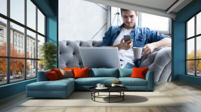 young man using his smartphone for online banking - sitting on sofa with laptop on leap. Wall mural