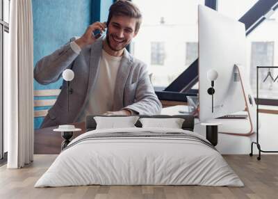 young smiling bearded businessman is sitting at table in front of computer, talking on cell phone. f Wall mural