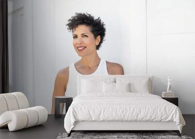 Smiling woman with dark short curly hair standing in front of a white wall in summer. Generative AI. Wall mural